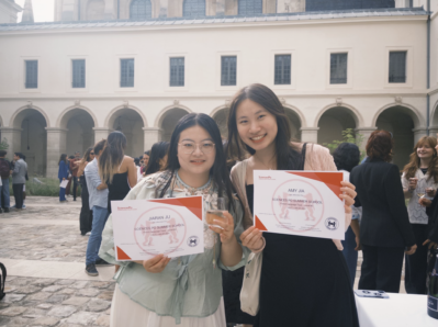 A couple of people holding certificates and drinks, standing in a courtyard with a stone walkway and an arched building in the background. Others are gathered socially in the area.